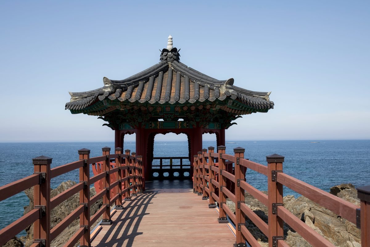 Traditional Korean wooden pavilion connected by a walkway over coastal rocks.