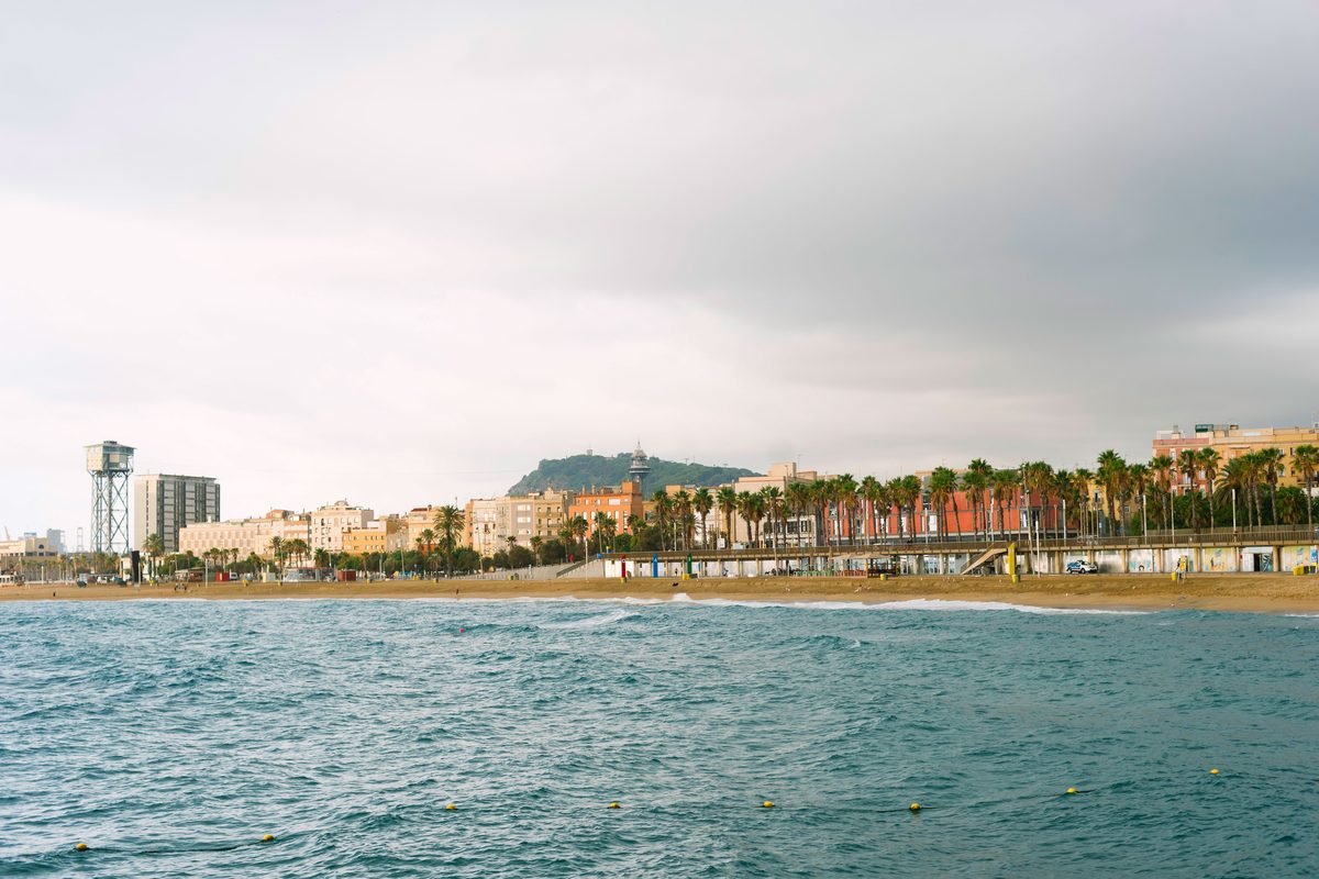 Coastal view of Barceloneta Beach with cityscape and palm trees under an overcast sky.