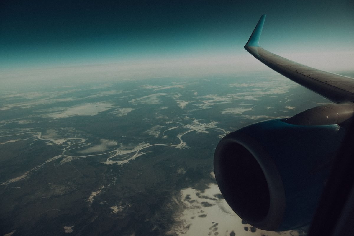 Through window of modern wing and turbine of airplane flying in sky above verdant land.