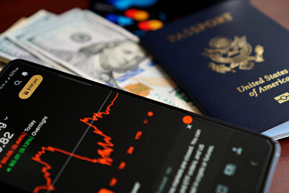 Flat lay of travel essentials: a passport, cash, and a smartphone displaying financial charts.