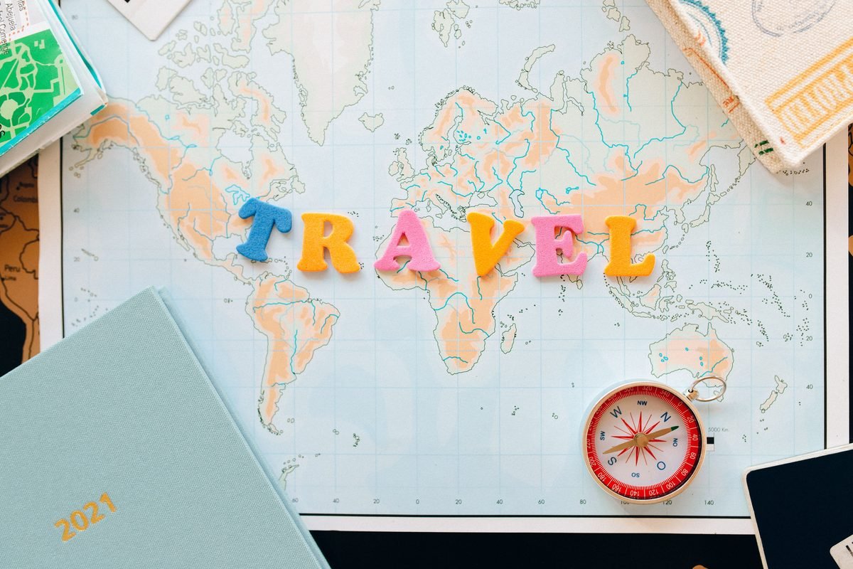 Flat lay of a world map with colorful letters spelling travel and a compass.
