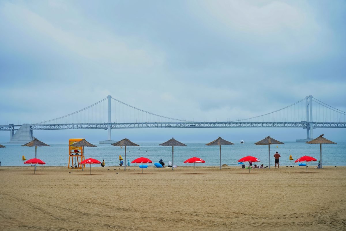 Sandy beach with colorful umbrellas and a long suspension bridge in the background