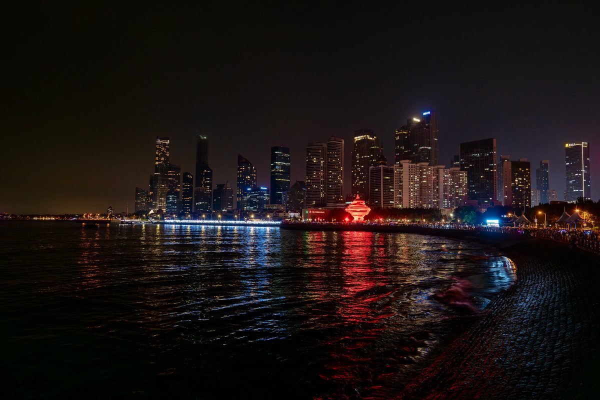 Illuminated skyscrapers reflecting on water along a dark shoreline at night