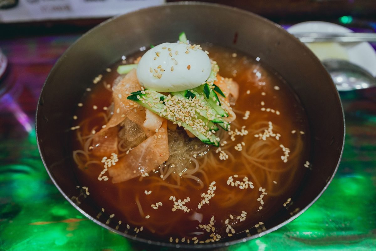 Metal bowl filled with Korean cold noodles garnished with cucumber and a boiled egg