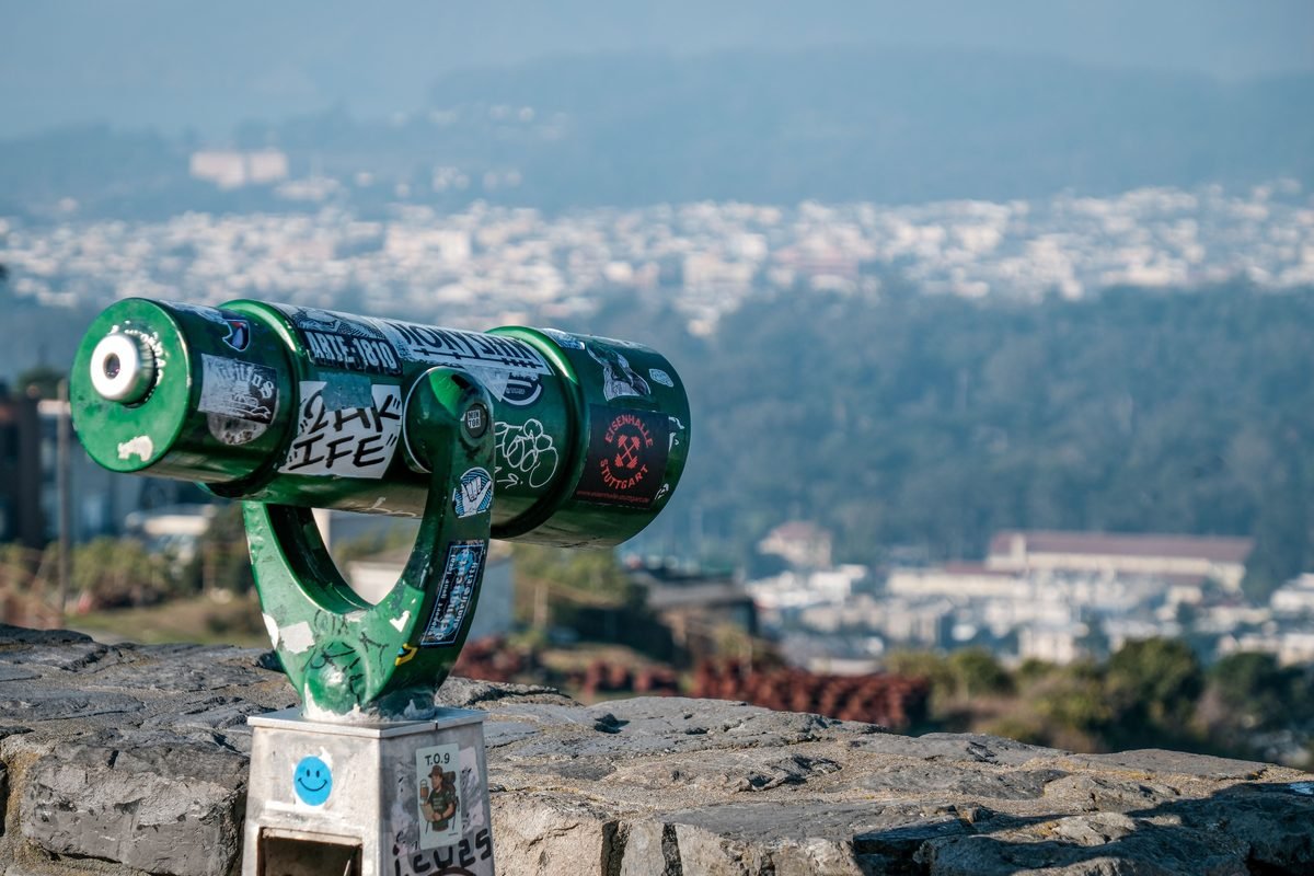 Green public binoculars overlooking a distant hazy cityscape.