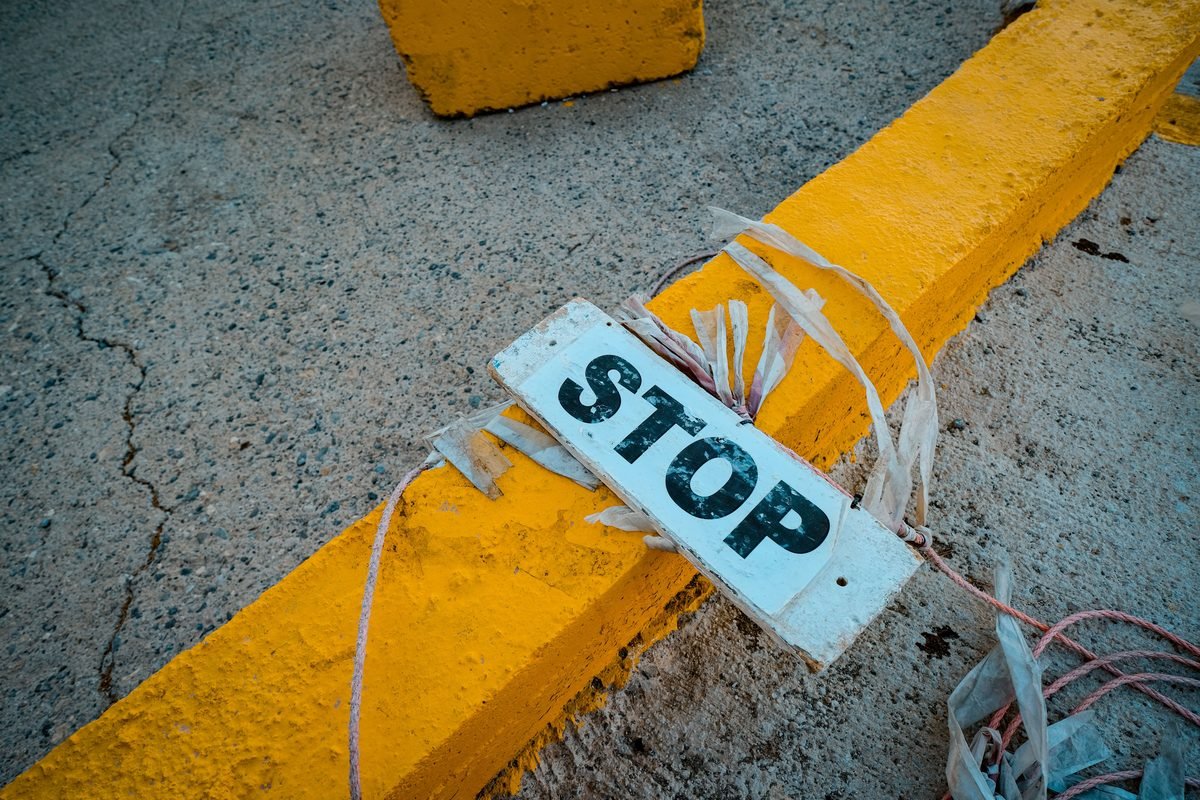 A worn stop sign placed on a cracked concrete pavement next to a yellow curb.