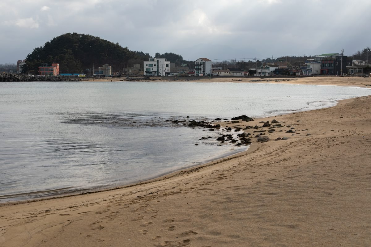 Sandy beach with calm ocean water and distant coastal buildings under cloudy skies.