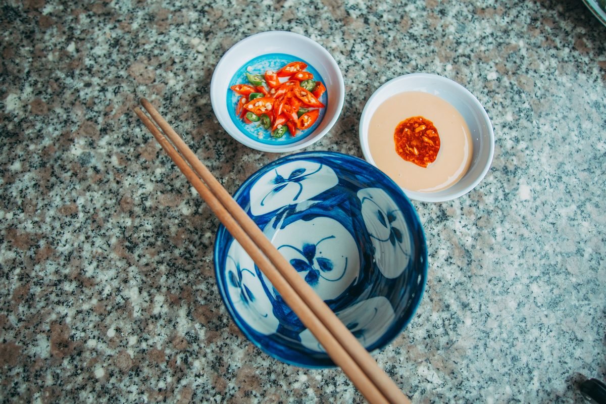 Small ceramic dishes of sliced red chili peppers and sauce with chopsticks on a stone table