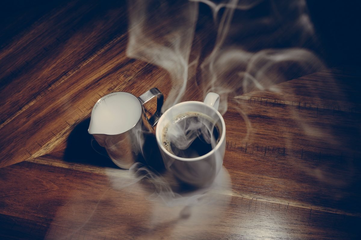 Steaming cup of black coffee with a milk jug on a wooden table.