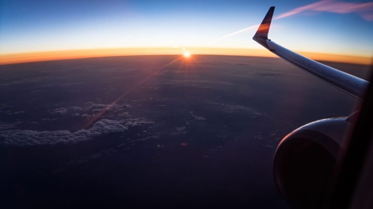 Aerial view from an airplane window showing the wing and a sunset over clouds.