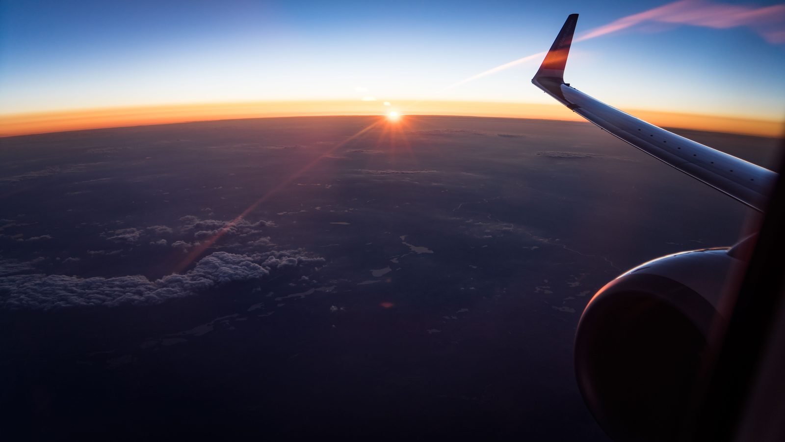 Aerial view from an airplane window showing the wing and a sunset over clouds.