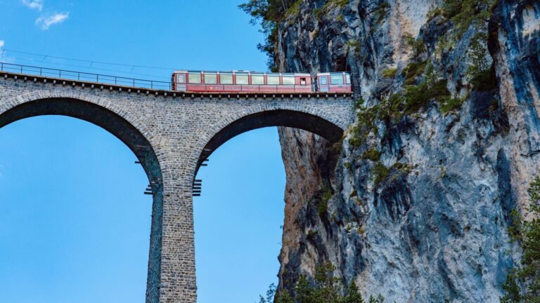 A red train crosses the Landwasser Viaduct against sheer rock faces in Switzerland.