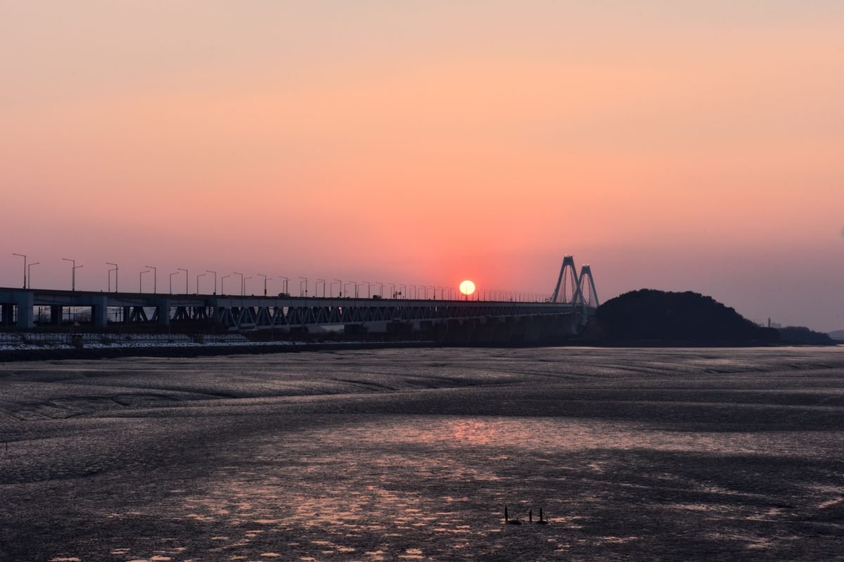 Yeongjong Bridge silhouetted against a warm orange sunset over tidal flats in Incheon.