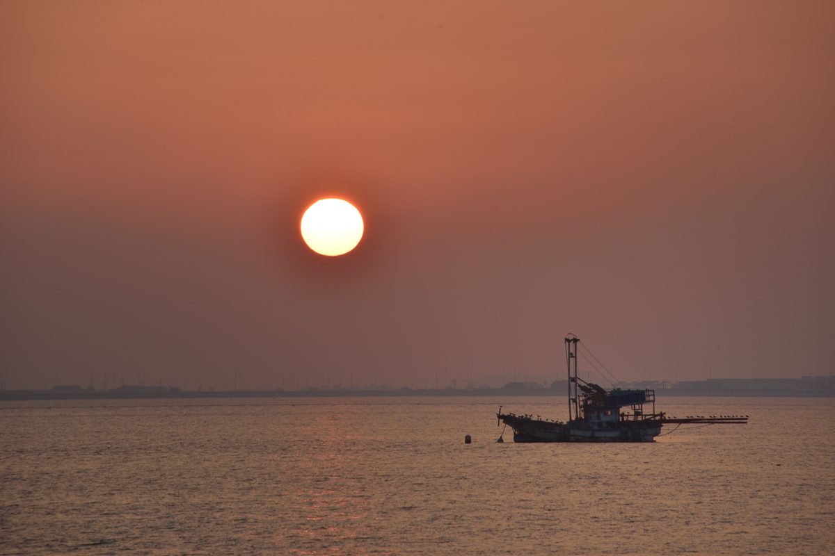 A fishing boat floating on calm water under a soft sunset sky with a bright round sun.