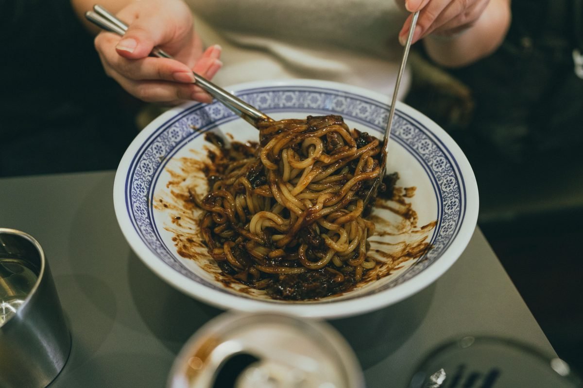 Person eating jjajangmyeon noodles with metal chopsticks from a traditional bowl.