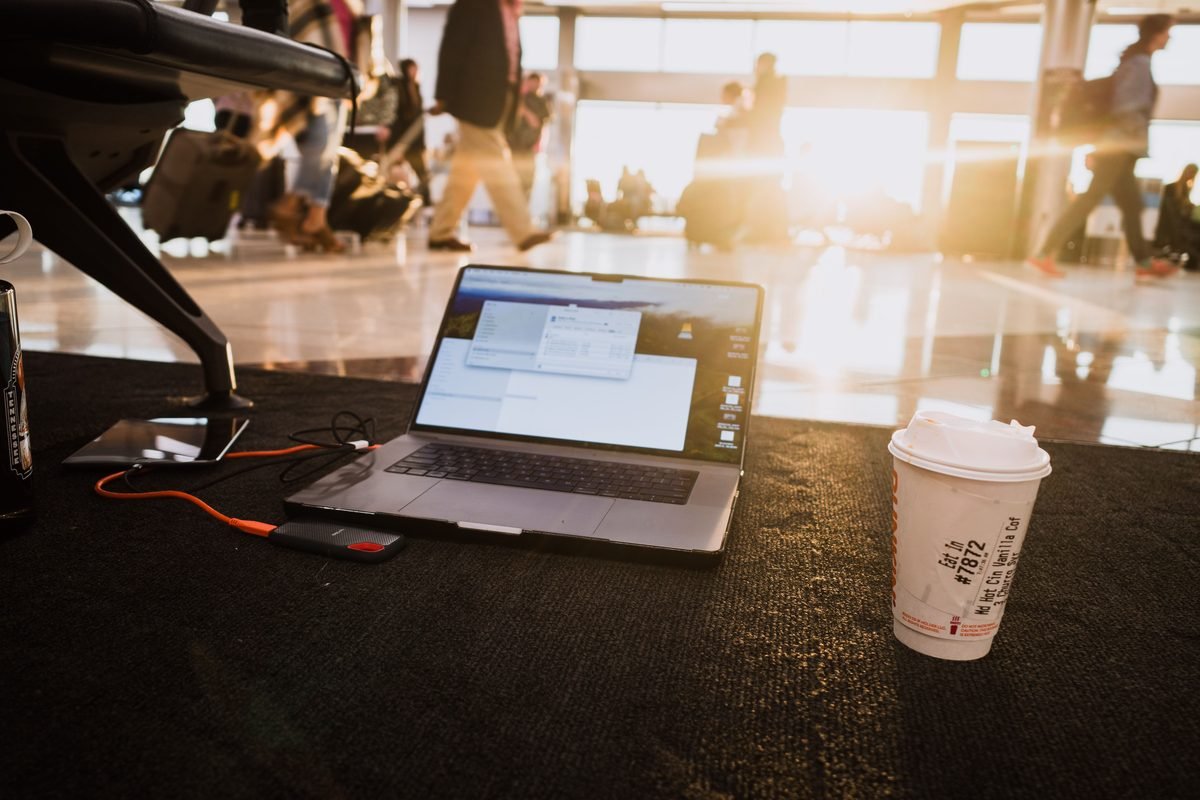 Laptop and coffee cup on airport floor with passengers.