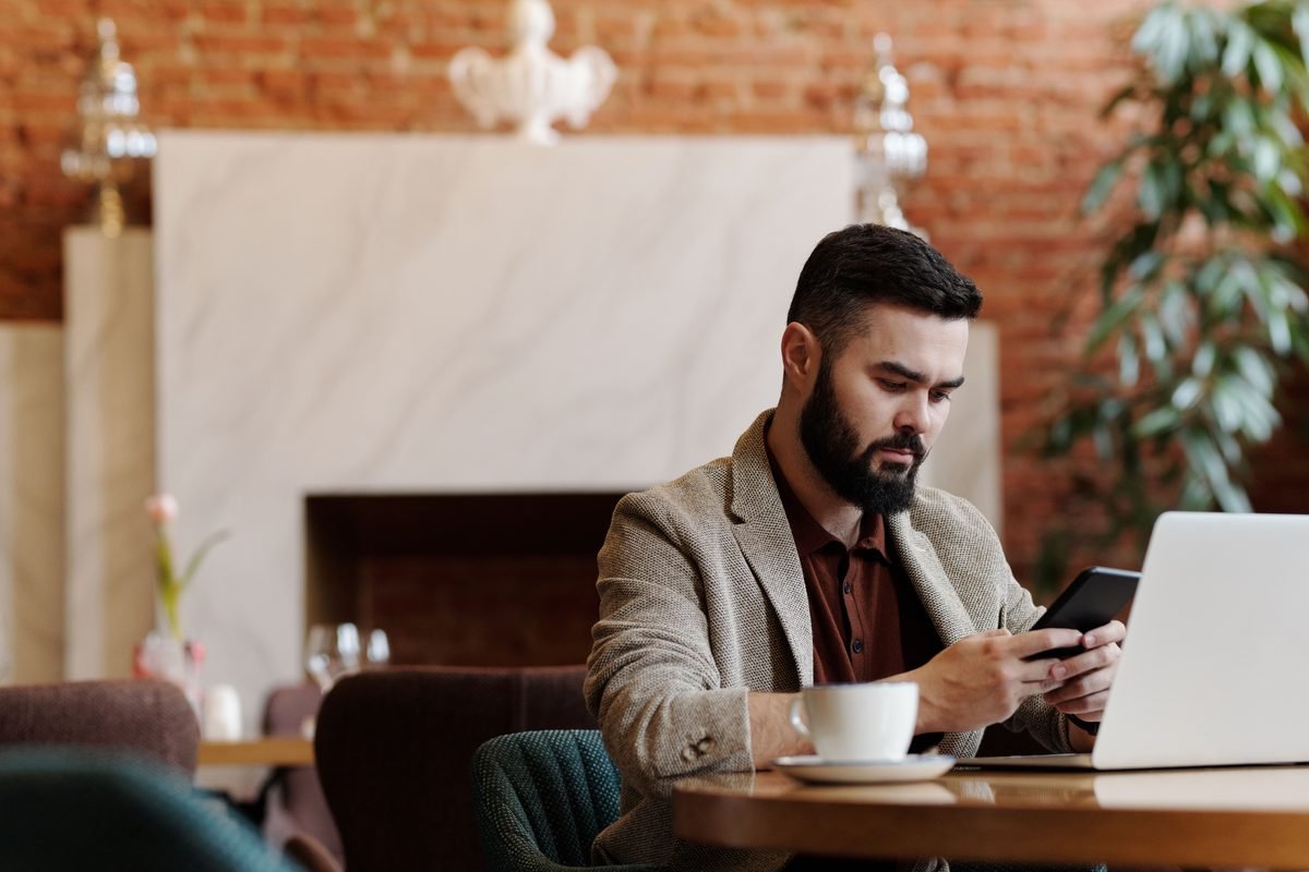 Man using smartphone and laptop for remote work in a cafe.