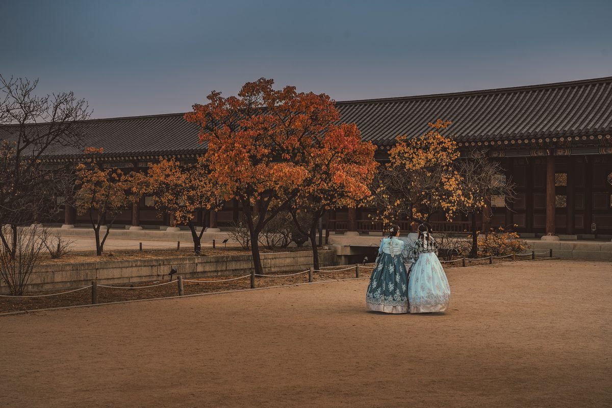 Two visitors in hanbok walk through Gyeongbokgung Palace in autumn.