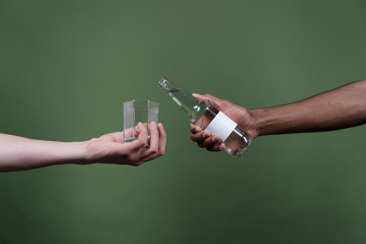 Hands pouring clear liquid from a clear bottle into a small glass.