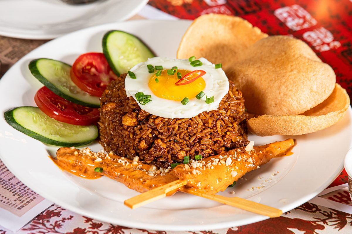 Plate of fried rice topped with a fried egg, cucumber, tomatoes, and satay skewers