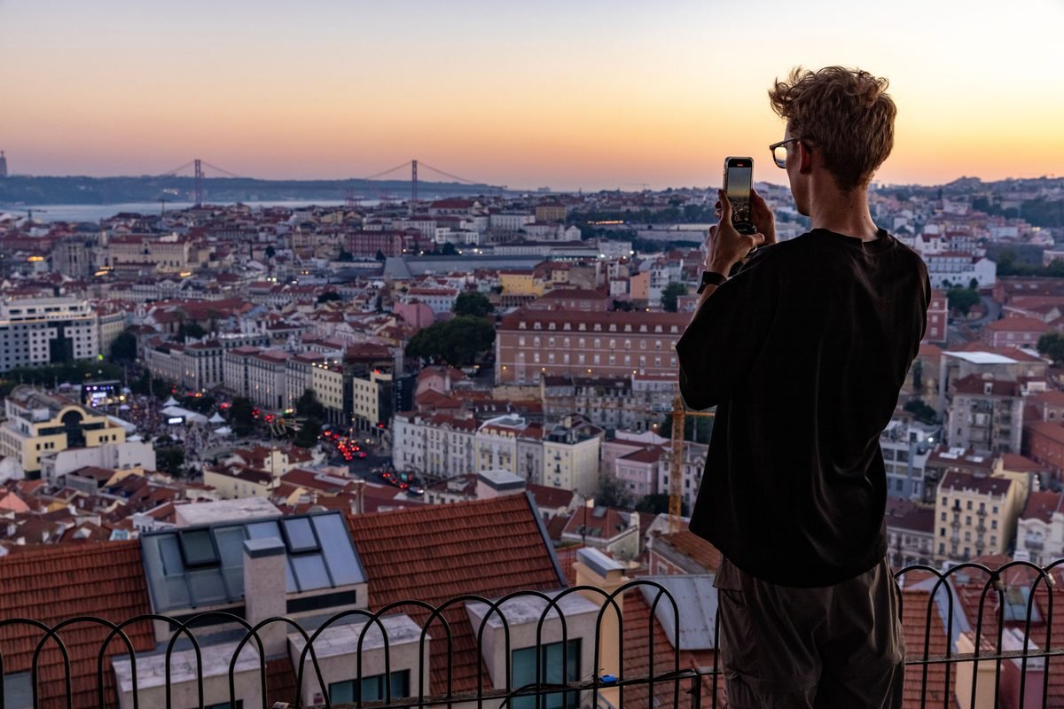 Tourist taking a photo of the golden hour sunset over the Lisbon cityscape from a viewpoint.
