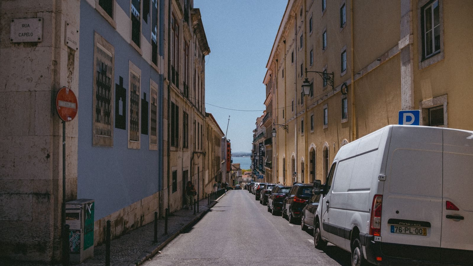 Historic street lined with buildings and parked vehicles leading toward the water in Lisbon.