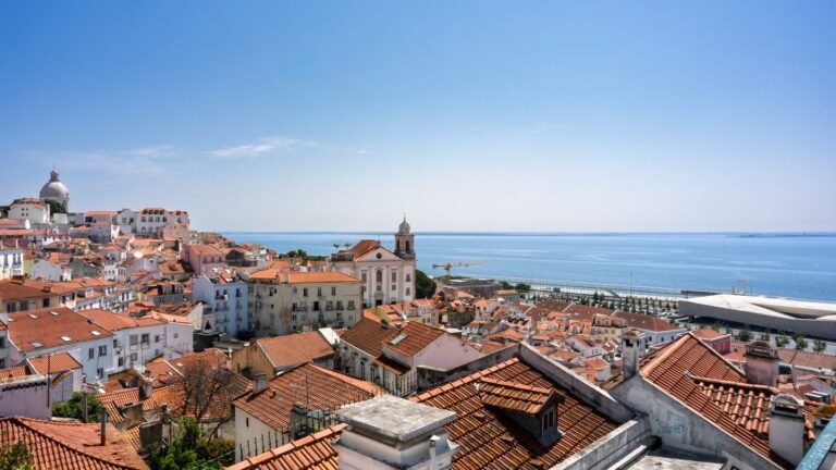 Panoramic view of Lisbon's terracotta rooftops cascading down toward the Tagus River under a blue sky.