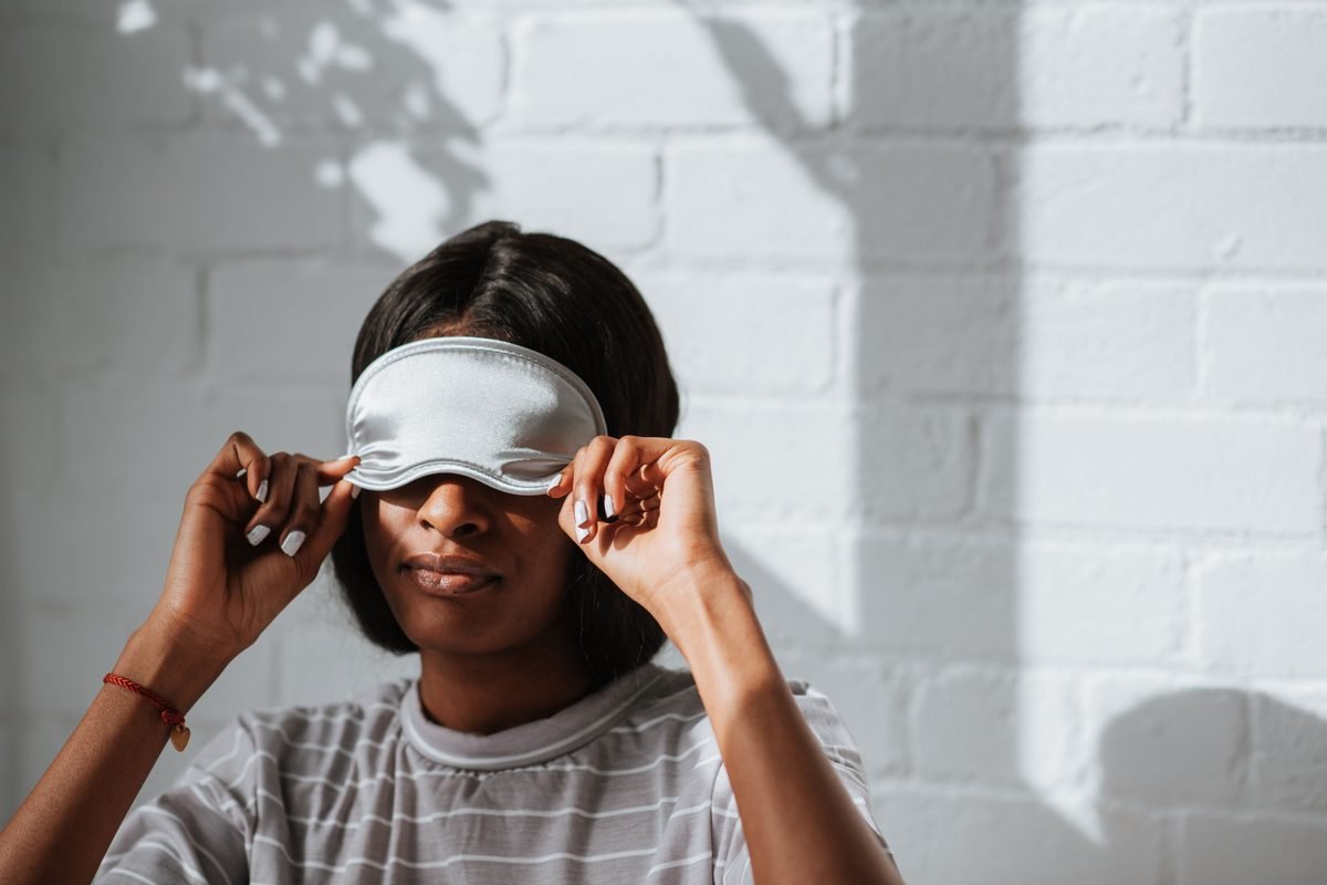 A woman wearing a grey striped shirt adjusting a sleeping mask indoors.