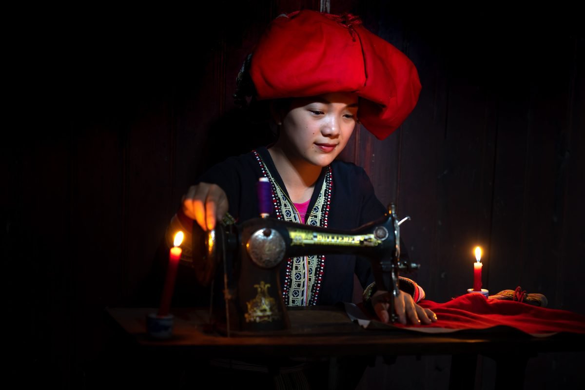Red Dao woman sewing by candlelight in traditional attire.
