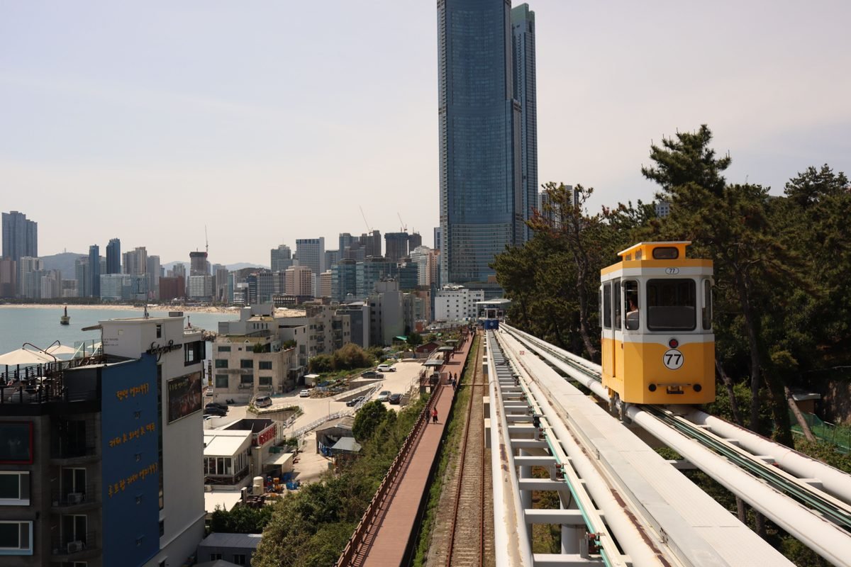 Yellow capsule train on elevated track overlooking skyscrapers and coast in Busan.