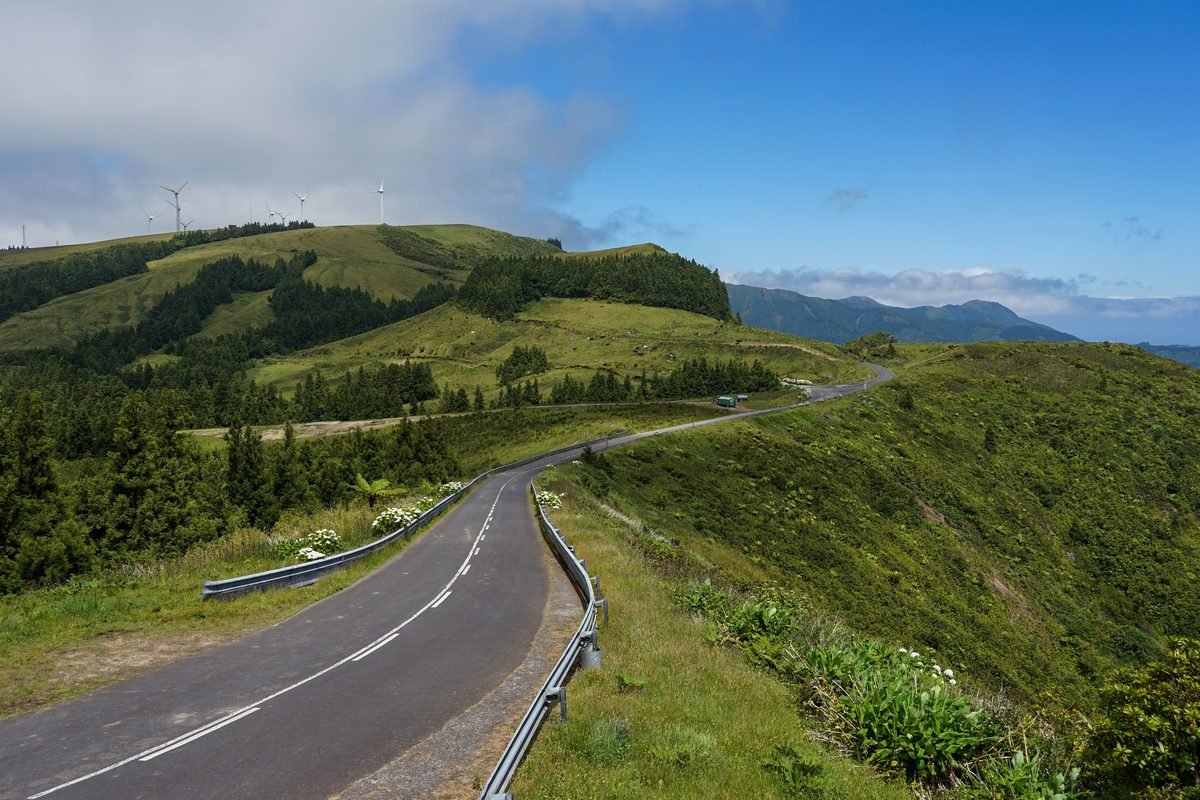 A winding road cuts through verdant hills with wind turbines under a blue sky.