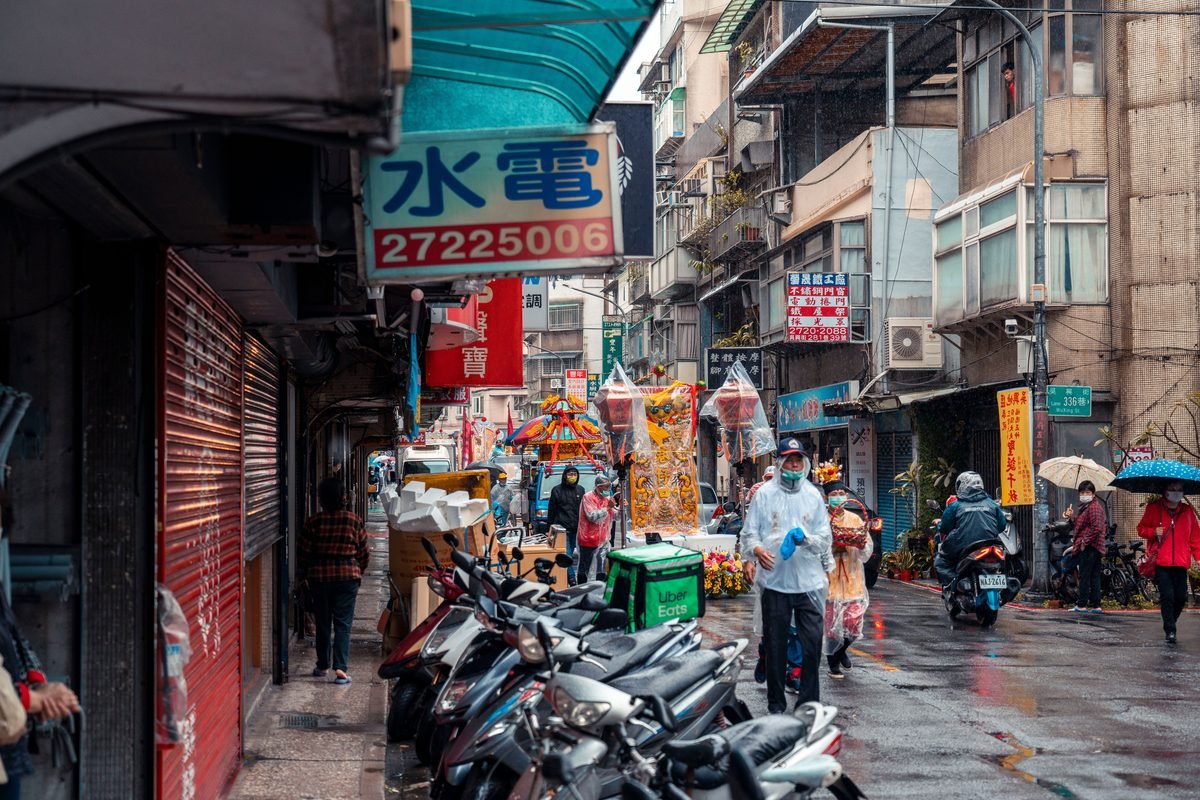 Narrow city street scene with scooters and pedestrians on a rainy day.