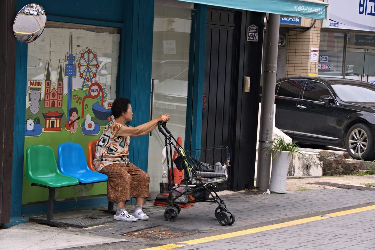 Elderly woman seated by a colorful mural on a street in Daegu