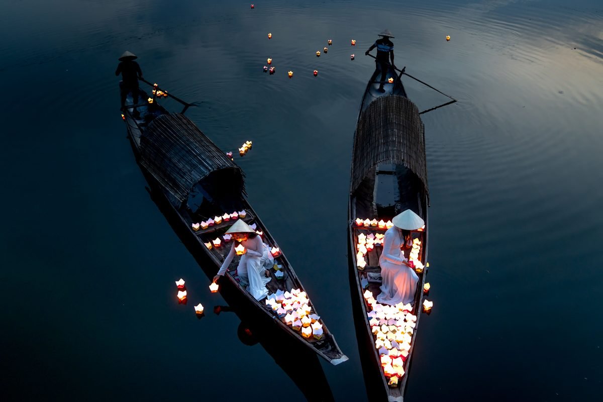 Boats carrying glowing lanterns on a river during twilight.
