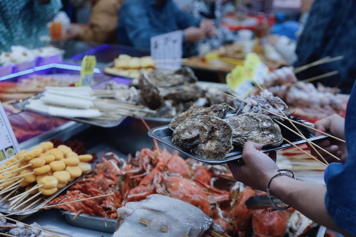 Hands holding a tray of grilled seafood skewers in a market stall