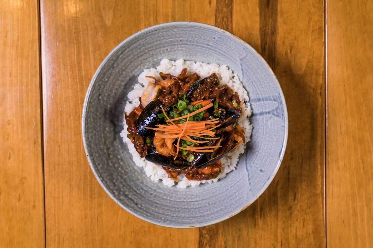 A ceramic bowl filled with rice topped with spicy seafood and sliced vegetables on a wooden table.