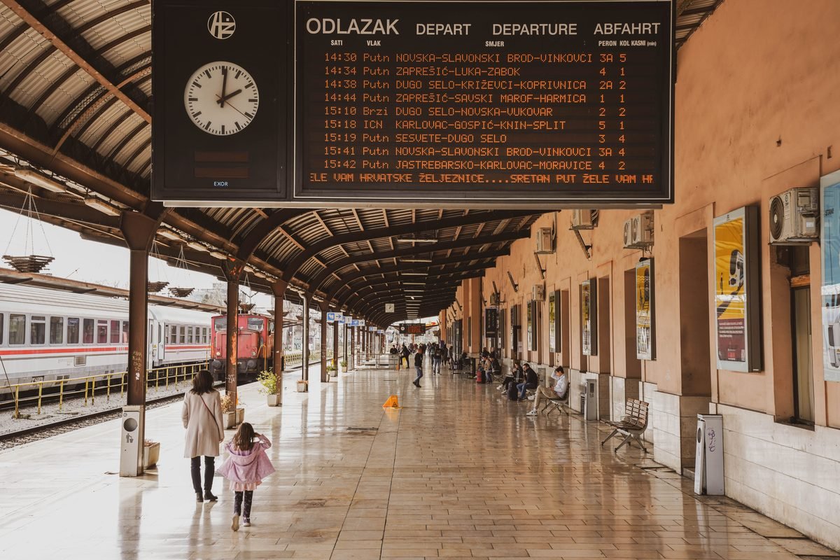 A train station platform with passengers and a large departure timetable display.
