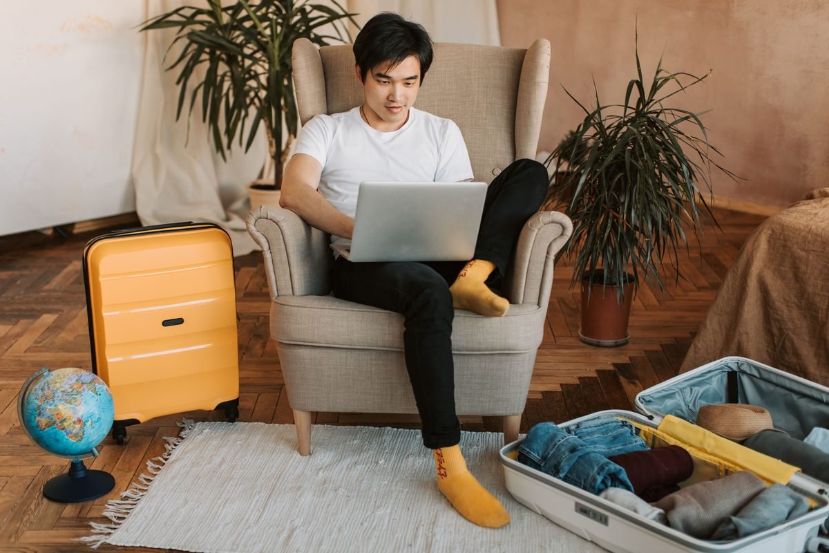 A man sitting in an armchair planning travel on a laptop with suitcases.