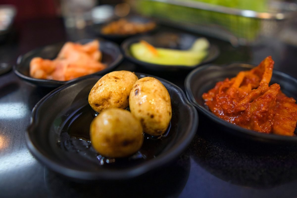 Close shot of Korean side dishes including baby potatoes and kimchi on small black plates.