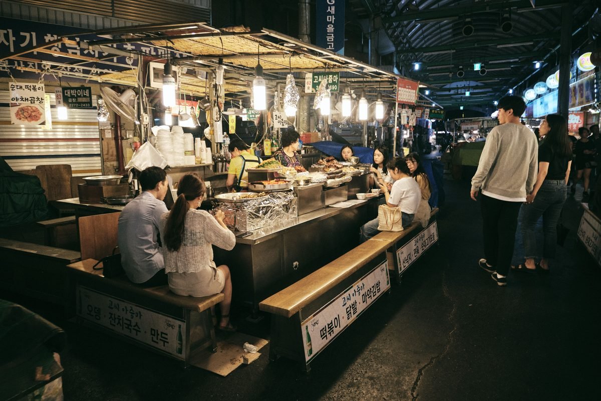 People seated and eating at an outdoor street food stall at night under bright lights.