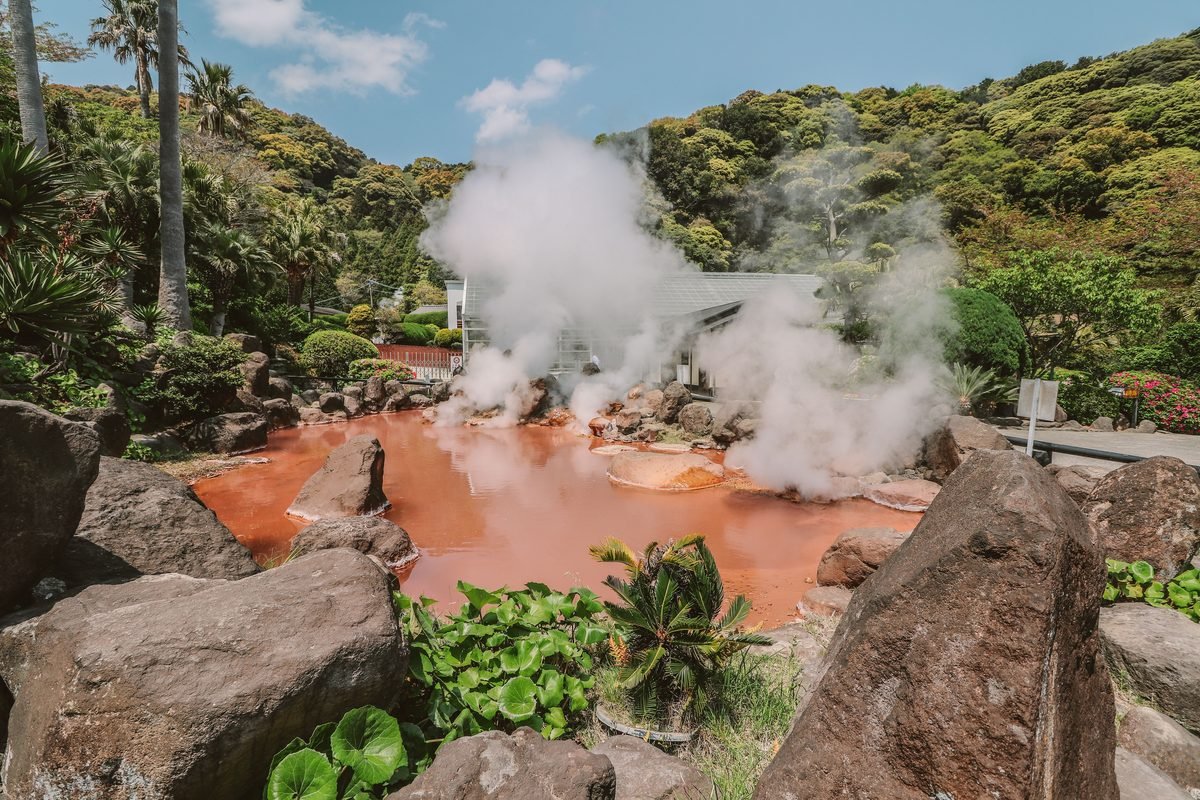 Vibrant reddish thermal spring pool emitting thick clouds of sulfur steam amid lush hills