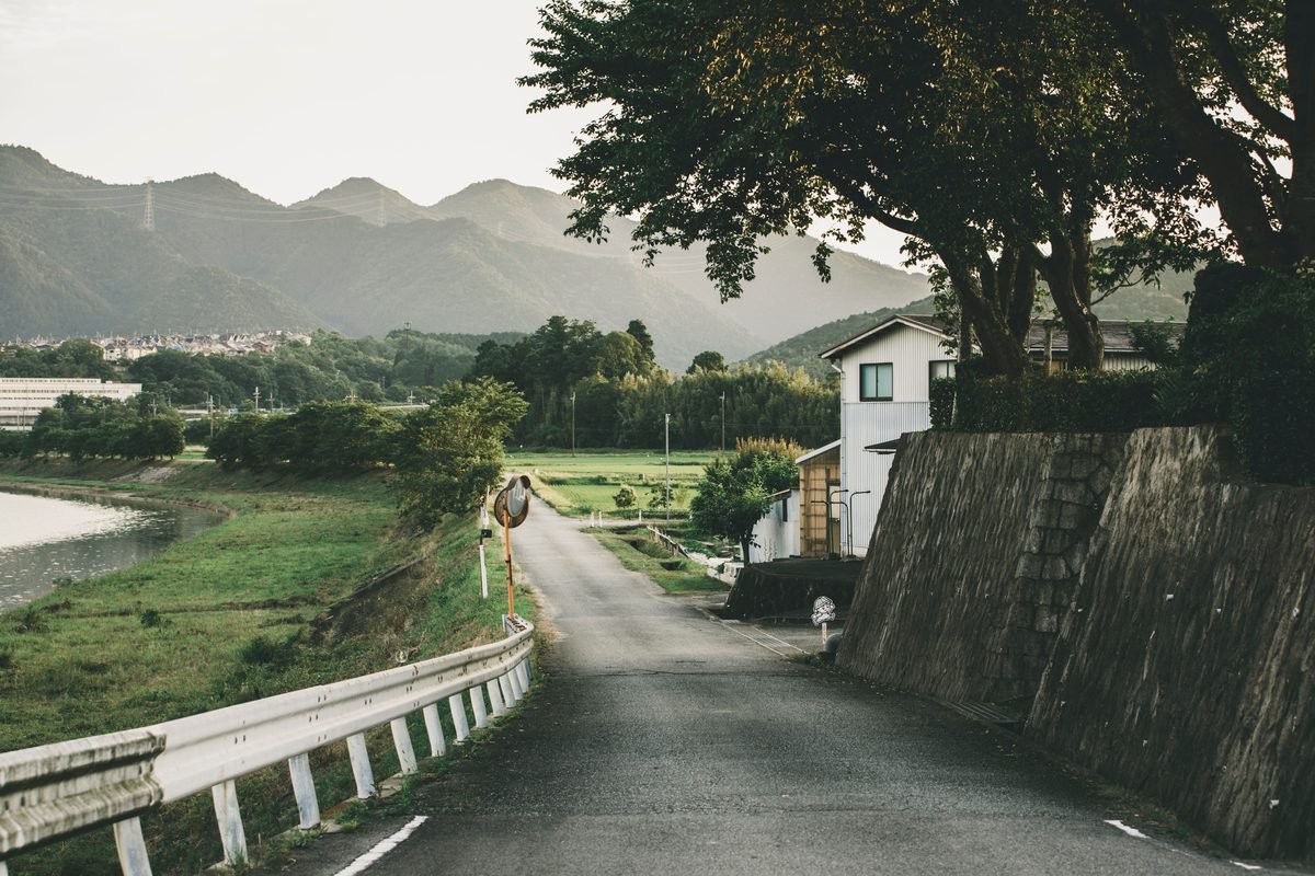 Quiet asphalt country road running alongside a grassy riverbank toward distant mountains