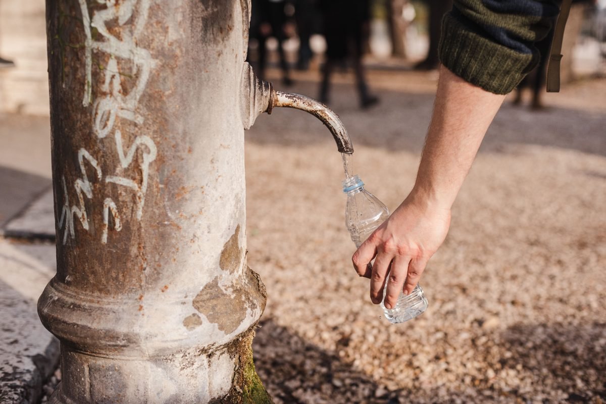Refilling a reusable water bottle at an old public fountain tap in a town