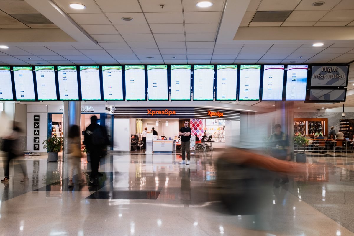 Travelers walking past a bank of large flight information screens in an airport terminal.