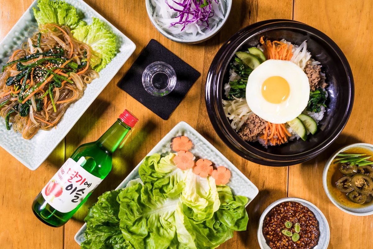 Korean feast featuring bibimbap, japchae, and a bottle of soju on a wooden table
