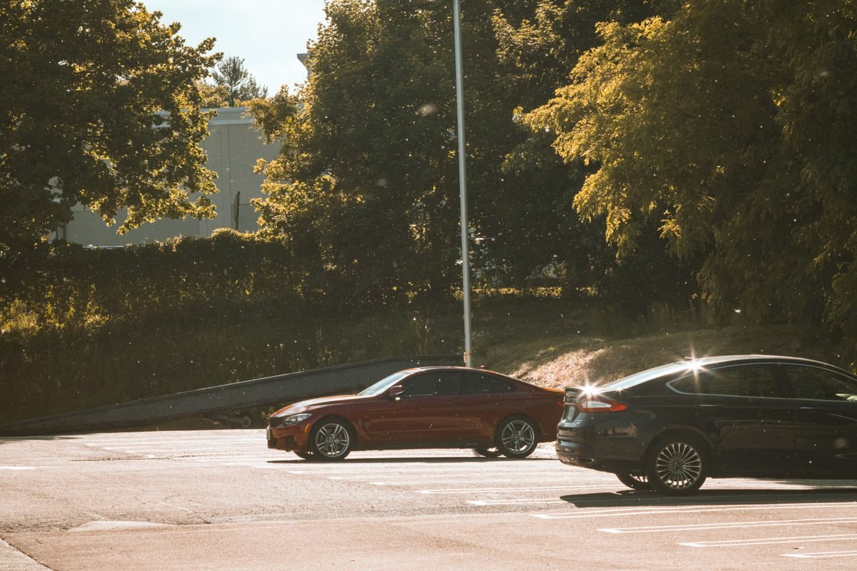 Cars parked under sunlit trees in an outdoor parking lot.