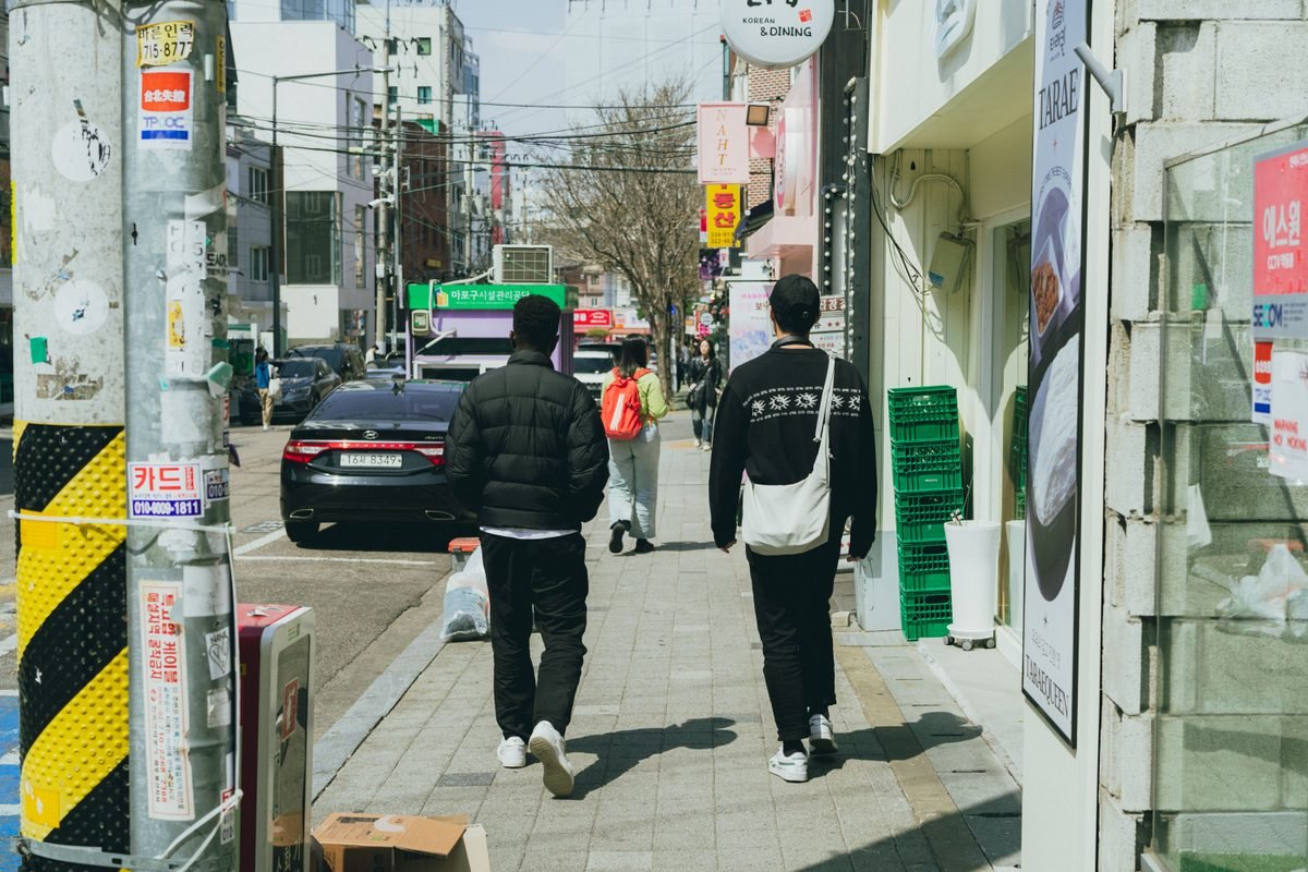 Pedestrians walking down a busy Seoul sidewalk lined with shops and utility poles.
