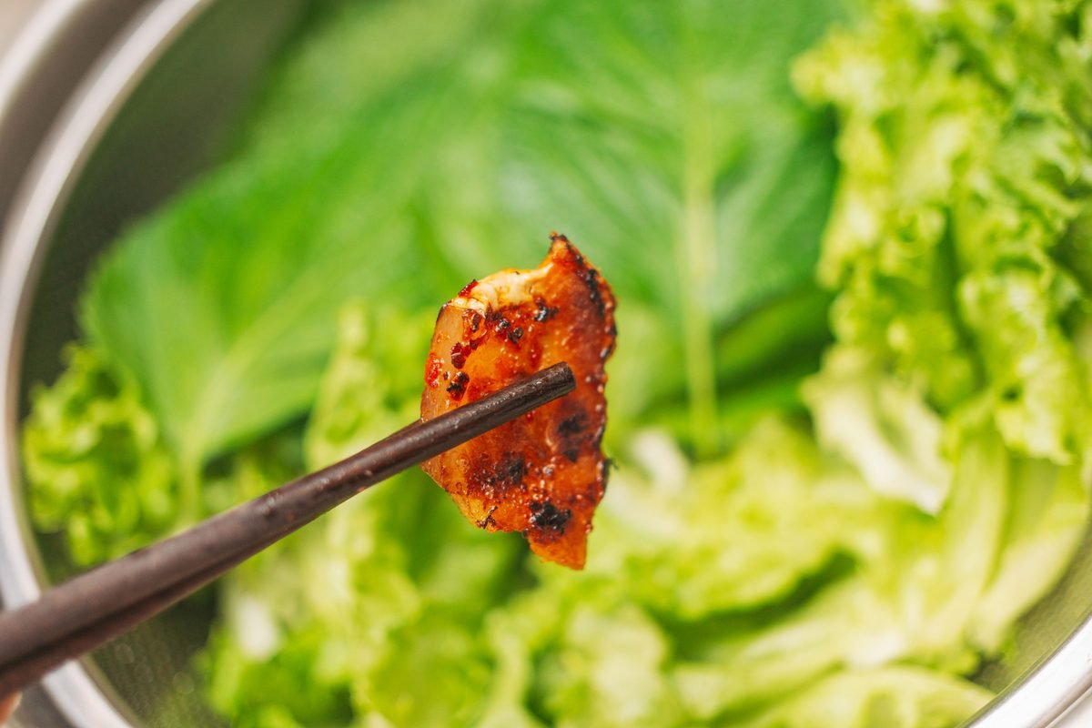 Grilled meat held by chopsticks over fresh leafy lettuce