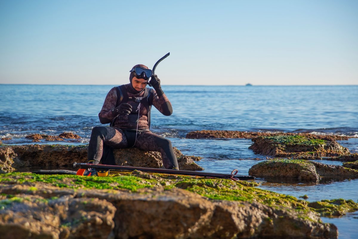 Man in wetsuit sitting on a rocky coastal shore preparing fishing equipment