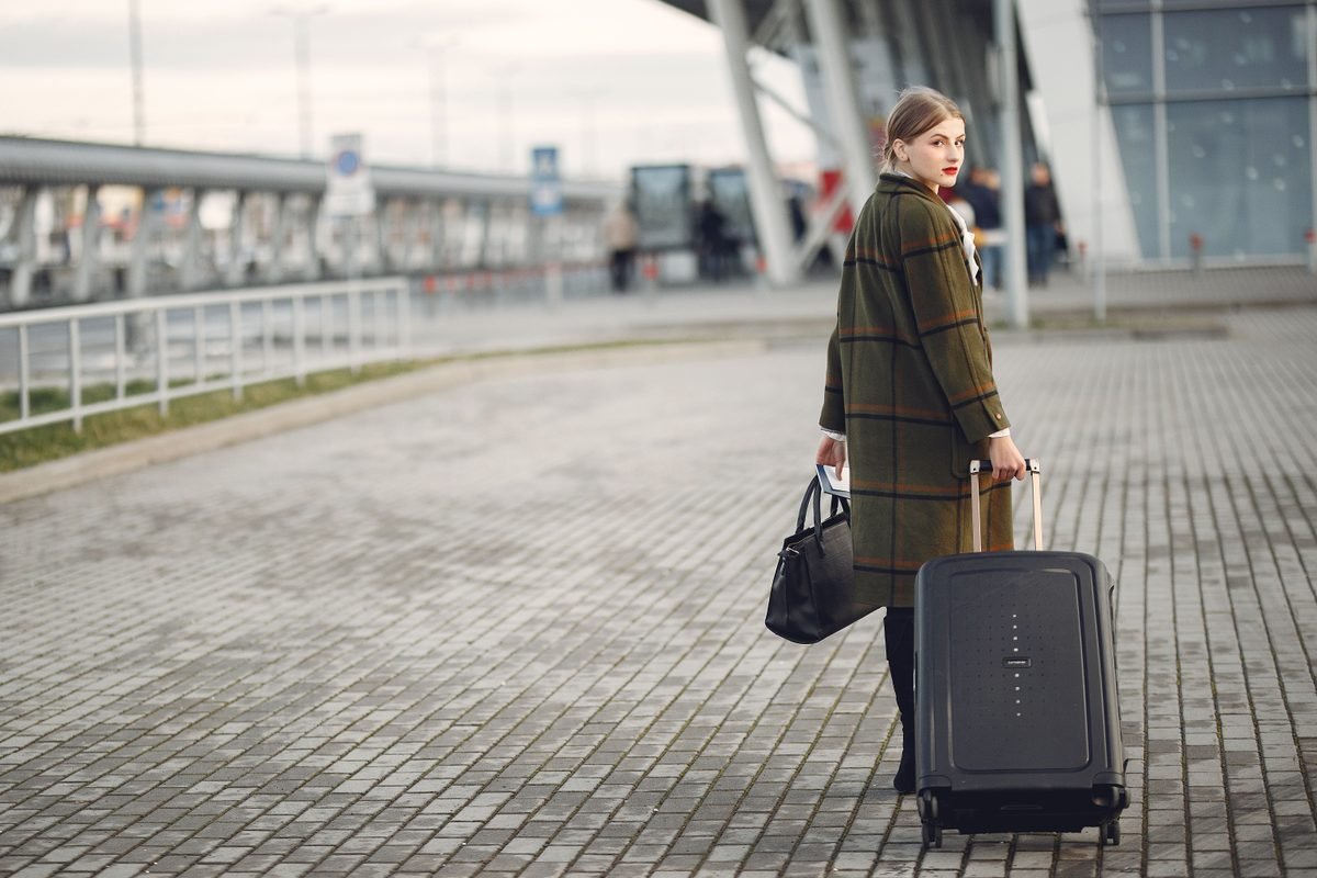 Stylish female traveler walking with suitcase outside airport terminal
